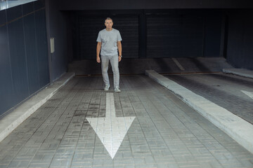 Full length of confident businessman standing on parking entrance on white arrow pointing to exit, gray automatic gates background. Underground car parking in modern office building