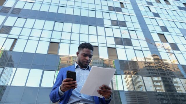 African American realtor checks information in paper document with phone. Man entrepreneur relates to contract of apartment purchase and sale with seriousness