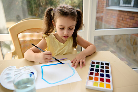 Adorable European Child, Elementary Age Smart Little Girl Using A Paintbrush, Painting Pictures With Watercolor Paints During An Art Class At Home. Kids Education And Entertainment. Hobby And Leisure