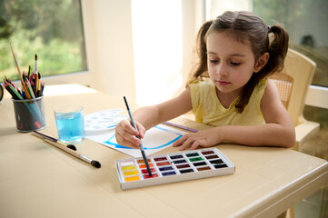 Talented little girl of preschool, holds paintbrush and mixes colorful paints on professional watercolor palette. Learning peinture. Art class. Kids education, hobby. Creative courses. Back to school