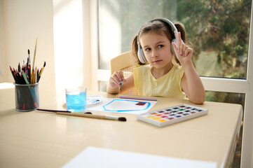 Charming talented European little girl of preschool wearing wireless headphones and yellow t-shirt, enjoying creative courses, learning watercolor painting. Art class. Hobby and leisure activity