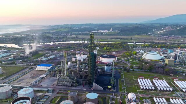Aerial orbit of a fractionation tower at an oil refinery in the middle of a natural landscape at sunset.