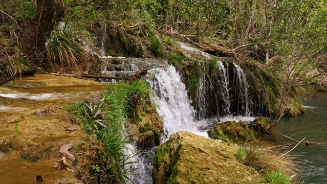 Small cascade of waterfall, green grass on stony edge, mediocre water flow falling down to little lake or pond. Bushes twigs seen on background. Turkey nature in details