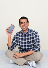 Asian man sitting in the floor smiling to camera while showing blank handphone screen