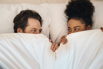 Interracial, happy and relaxed couple lying in bed, bonding and looking shy while hiding after...