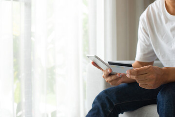 Young man wearing white t-shirt sitting on sofa in the living room and hand holding credit card and smart phone. He uses his credit card for online purchases via his mobile phone.
