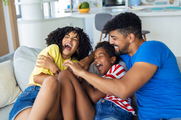 Cheerful people sitting on couch in living room have fun little daughter tickling mother laughing...