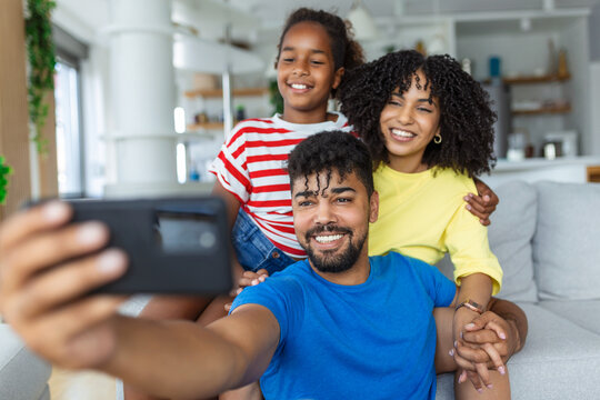 Cheerful family vloggers with child daughter taking selfie, recording vlog, making video call on phone having fun together in living room, parents and kid girl laugh using cellphone funny online app