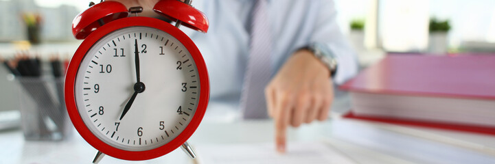 Man hand on red alarm clock stands at desk in office showing seven o'clock AM PM