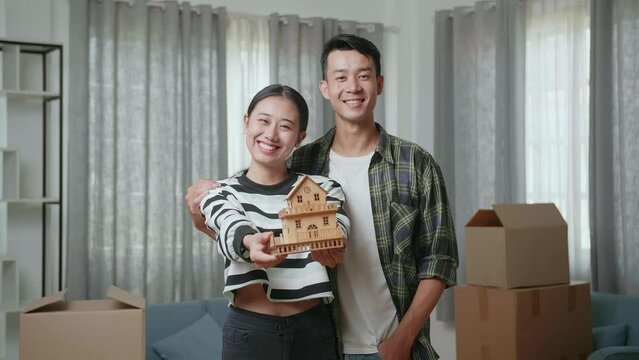 Young Asian Couple With Cardboard Boxes On The Floor Smiling And Showing House Model To Camera In The New House
