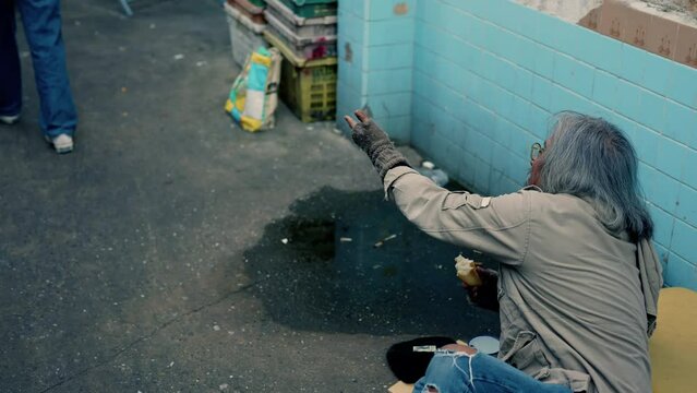 4k, An old Asian man, a homeless man dressed raggedly with long hair, sits eating bread in a narrow alley, begging people for food and money. Homeless people who are homeless have to sleep on streets