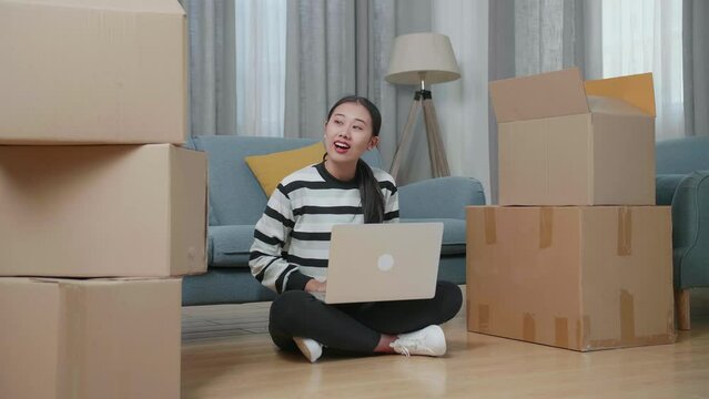 Young Asian Woman With Cardboard Boxes Sitting On The Floor And Comparing The House To The Photo On A Laptop After Moving Into A New House 
