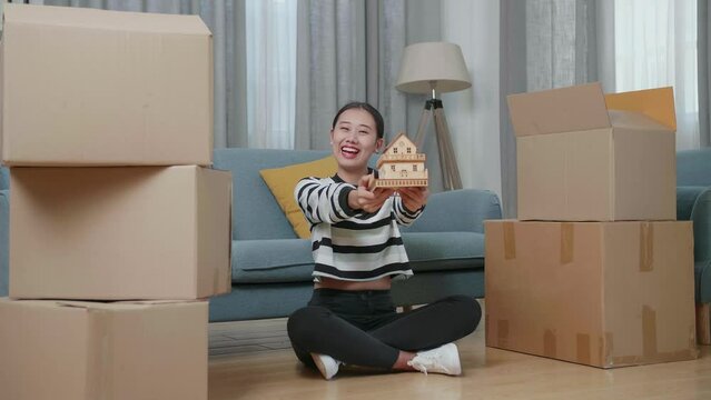 Young Asian Woman With Cardboard Boxes Sit On The Floor Smiling And Showing House Model To Camera In The New House
