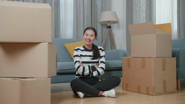 Young Asian Woman With Cardboard Boxes Sit On The Floor Smiling And Crossing Her Arms To Camera In The New House
