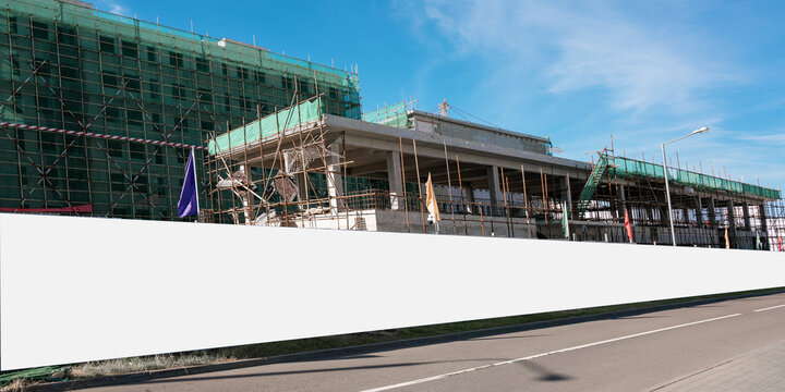 Long blank hoarding with white canvas mounted on construction site with unfinished building with wall and scaffold