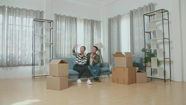 Young Asian Couple With Cardboard Boxes On The Floor Sitting On A Sofa And Taking Photo By Smartphone After Moving Into A New House 
