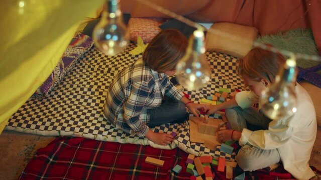 A zooming in shot of two boys focusing on building a tower together with blocks while sitting down in a luminous indoor sheet fort