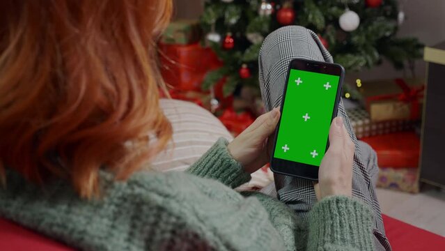 A red haired woman close up tapping and scrolling on mobile phone in Christmas decorated living-room. A girl browsing online.