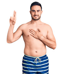 Fototapeta premium Young handsome man wearing swimwear smiling swearing with hand on chest and fingers up, making a loyalty promise oath