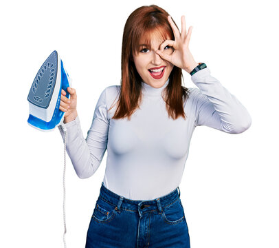 Redhead young woman holding electric steam iron smiling happy doing ok sign with hand on eye looking through fingers