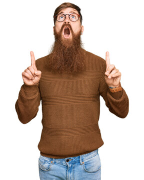 Young irish redhead man wearing casual clothes and glasses amazed and surprised looking up and pointing with fingers and raised arms.
