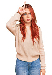 Young redhead woman wearing casual winter sweater making fun of people with fingers on forehead...