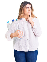 Young caucasian woman holding recycling plastic bottles serious face thinking about question with hand on chin, thoughtful about confusing idea