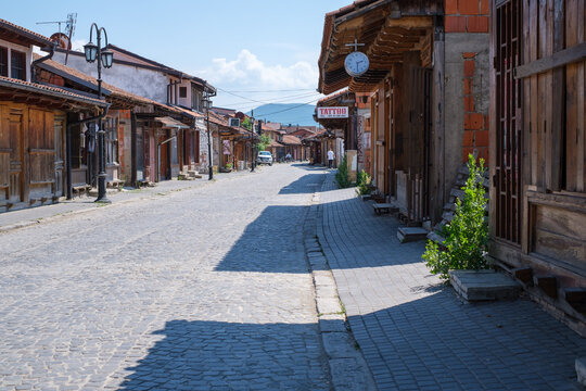 A narrow cobblestone street in Gjakova, Kosovo, lined with traditional wooden shops and houses under a bright blue sky. Perfect for travel, culture, and architecture themes.