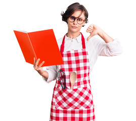 Beautiful young woman with short hair wearing professional baker apron reading cooking recipe book with angry face, negative sign showing dislike with thumbs down, rejection concept