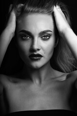 Fototapeta premium Fashion concept. Close-up black and white studio portrait of beautiful woman with long bright hair looking at camera with seductive look. Model holding hands over head and brushing hair with fingers