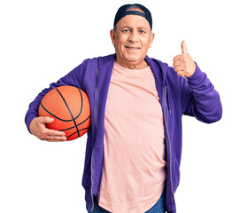 Senior handsome grey-haired man holding basketball ball smiling happy and positive, thumb up doing...