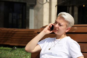 Girl with short blonde hair talking on mobile phone while sitting on street wooden bench. Tomboy...