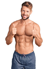 Young caucasian man standing shirtless success sign doing positive gesture with hand, thumbs up smiling and happy. cheerful expression and winner gesture. - obrazy, fototapety, plakaty
