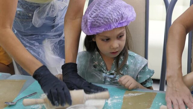 Pretty 5-year old girl rolls out raw dough with a small rolling pin on a silicone mat at the master class. Preparing dough for baking pizza, pie. Stages of baking and confectionery preparation.