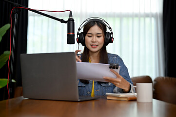 Charming Asian woman recording a podcast on her laptop with headphones and a microphone.