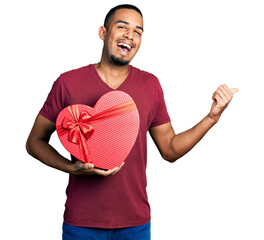 Young african american man holding valentine gift pointing thumb up to the side smiling happy with...