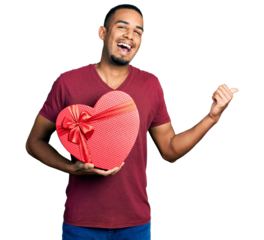 Young african american man holding valentine gift pointing thumb up to the side smiling happy with open mouth