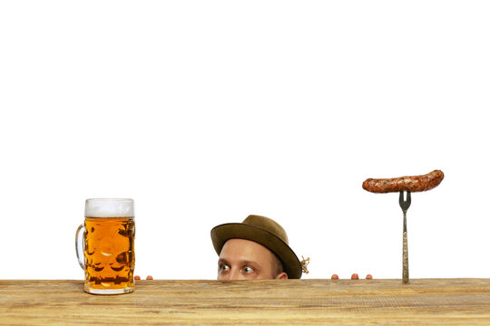 Portrait Of Young Man Peeking Out Table And Looking At Mug With Foamy Lager Beer And Sausage Isolated Over White Background