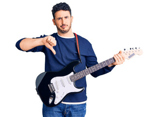 Young hispanic man playing electric guitar with angry face, negative sign showing dislike with thumbs down, rejection concept