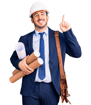 Young hispanic man wearing architect hardhat and leather bag holding blueprints surprised with an idea or question pointing finger with happy face, number one