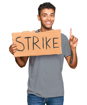 Young handsome african american man holding strike banner cardboard surprised with an idea or question pointing finger with happy face, number one