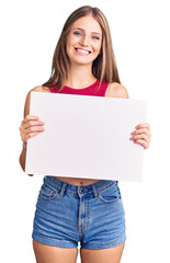 Fototapeta premium Young beautiful blonde woman holding blank empty banner looking positive and happy standing and smiling with a confident smile showing teeth