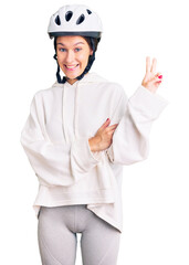 Beautiful brunette young woman wearing bike helmet and sporty clothes smiling with happy face winking at the camera doing victory sign. number two.