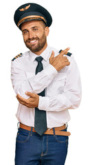 Handsome man with beard wearing airplane pilot uniform with a big smile on face, pointing with hand and finger to the side looking at the camera.