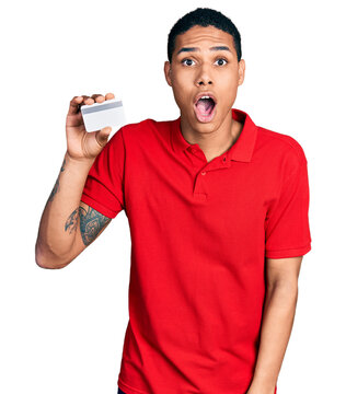 Young hispanic man holding credit card scared and amazed with open mouth for surprise, disbelief face