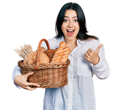 Beautiful hispanic woman holding wicker basket with bread pointing thumb up to the side smiling happy with open mouth