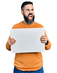 Hispanic man with beard holding blank empty banner angry and mad screaming frustrated and furious, shouting with anger. rage and aggressive concept.