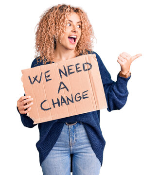 Young blonde woman with curly hair holding we need a change banner pointing thumb up to the side smiling happy with open mouth