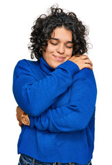 Young hispanic woman with curly hair wearing turtleneck sweater hugging oneself happy and positive,...