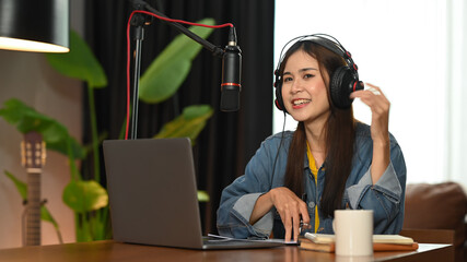 Woman podcaster wearing headphone talking into a microphone while making an online podcast from her...